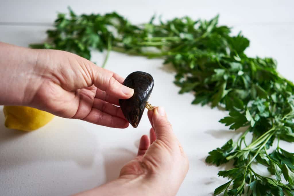 A woman's hands are shown pulling the beard out of a mussel. Parsley and lemon are in the background on a white surface.