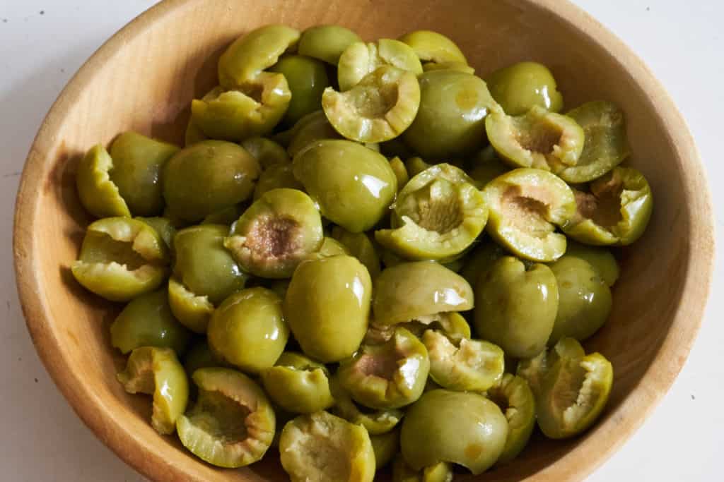 A wooden bowl full of Castelvetrano olives that have been torn in half.