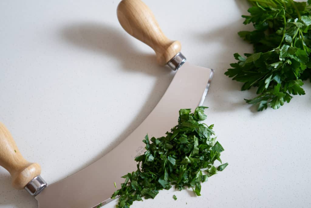 A mezzaluna with wooden handles, chopped parsley, and a bundle of fresh parsley on a white surface.