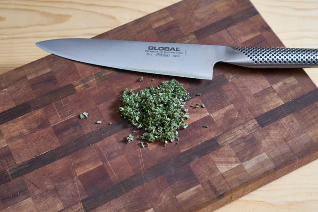 A chef's knife on a wooden cutting board beside chopped fresh sage.
