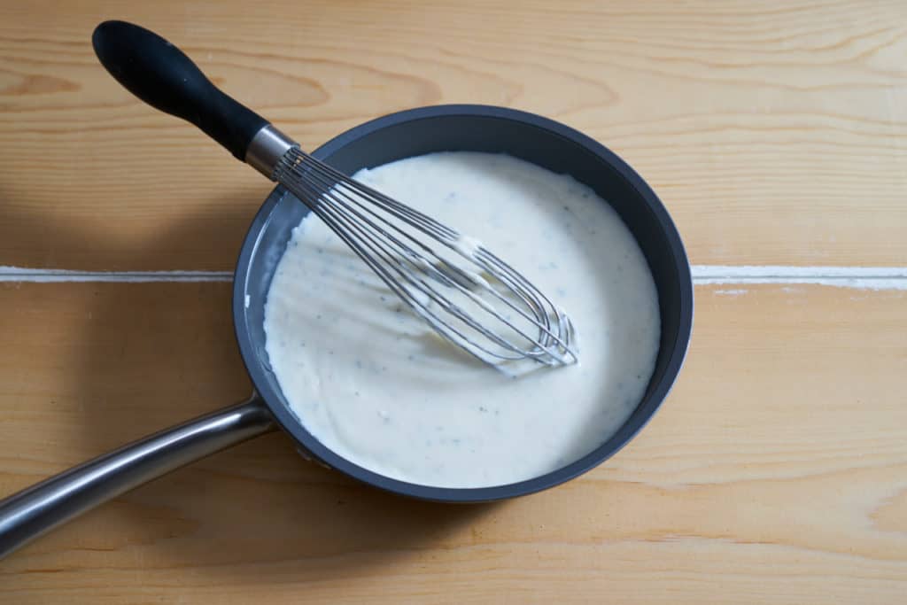 Bechamel sauce with fresh sage in a small skillet on a wooden surface. A whisk is in the skillet resting on the left side.