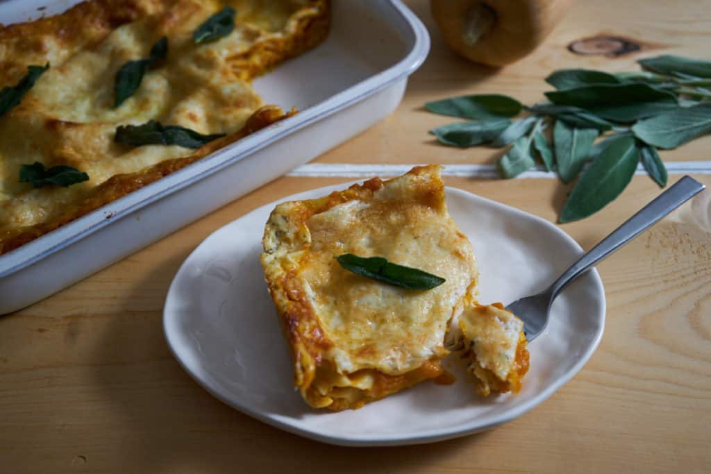 A portion of butternut squash lasagna with a fork on a white plate. A white baking dish filled with the lasagna and fresh sage are in the background.