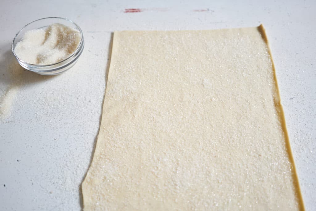 A sheet of puff pastry covered in sugar on a white surface. A bowl of sugar sits on the left. 