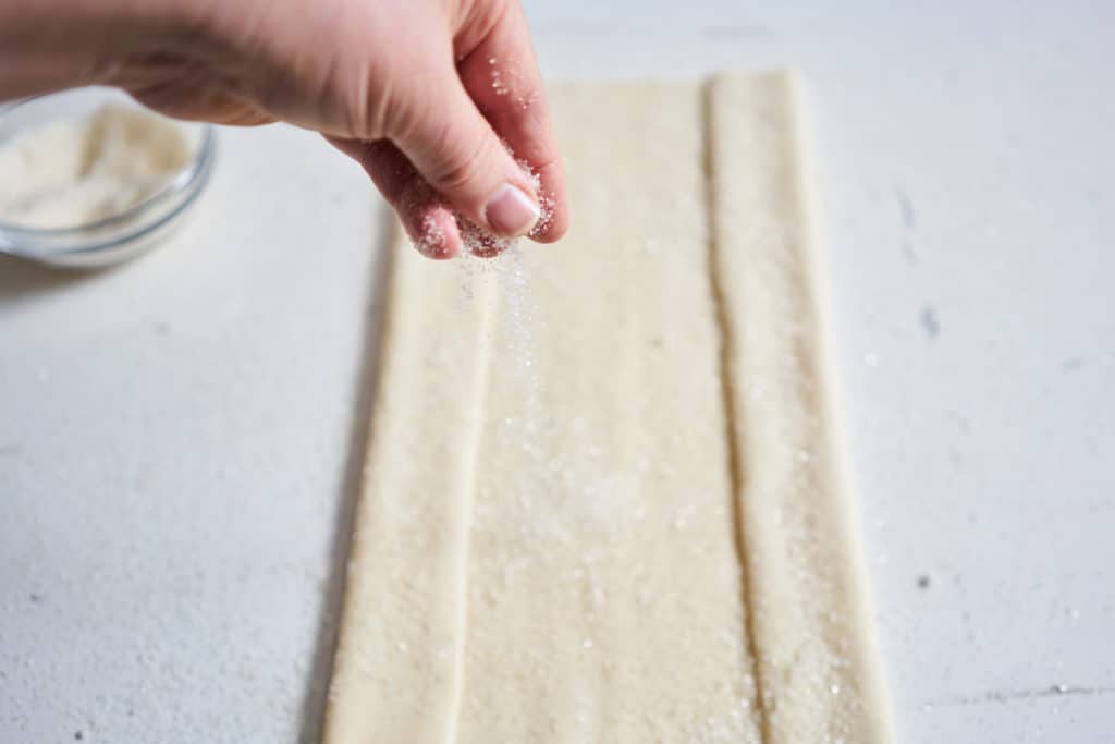 A woman's hand is sprinkling sugar onto a sheet of puff pastry that has been folded at the edges.