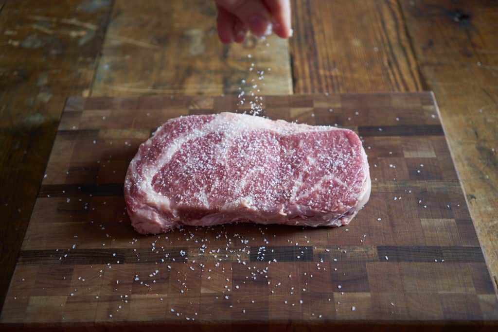 A woman's hand is shown seasoning raw ribeye steak with salt.