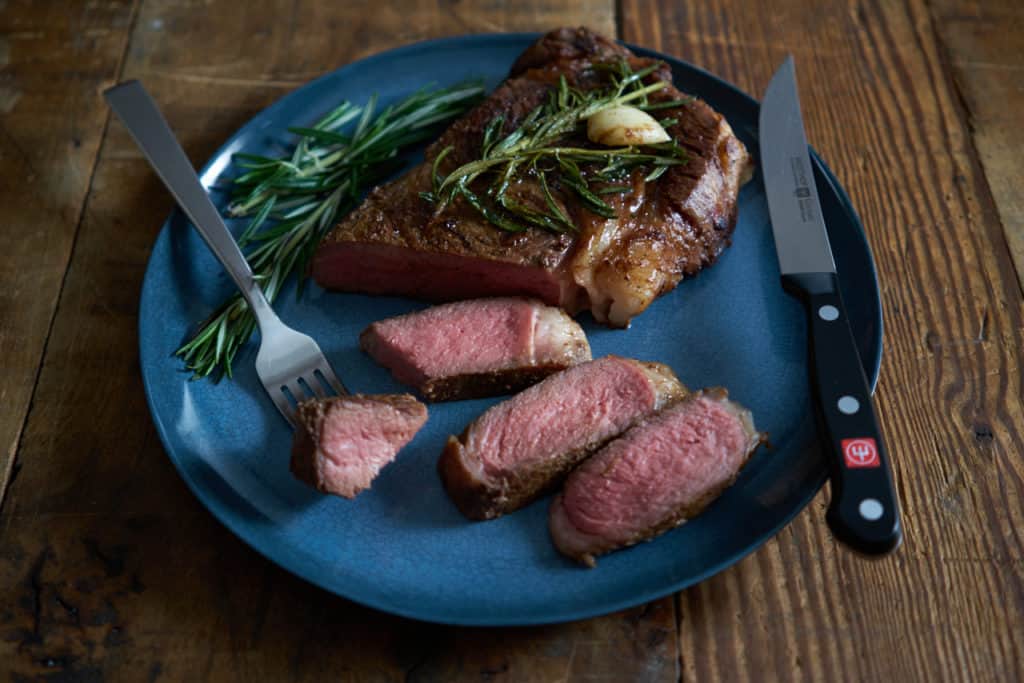 A blue plate on a wooden surface with a knife and fork and a sliced ribeye steak cooked to medium rare that is pink all the way through, garnished with rosemary and garlic.
