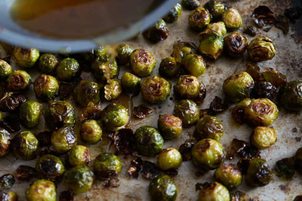 Honey-lime syrup is being poured from a skillet onto roasted brussels sprouts on a baking sheet.