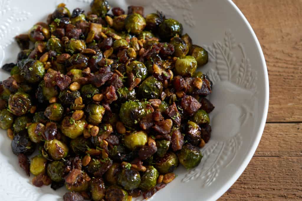 Roasted brussels sprouts with pistachios, dates, and lime in a white serving bowl on a wooden surface.