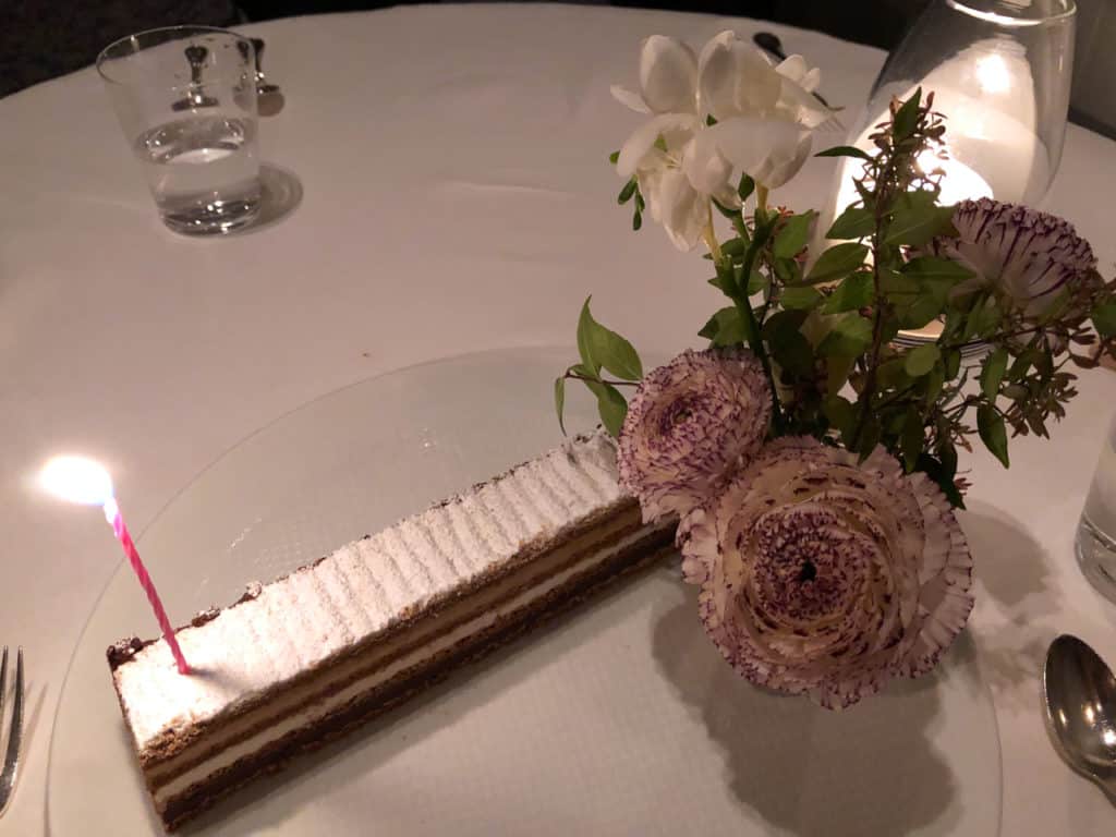 A long, rectangular cake with a candle on a white plate. White and purple flowers on on the right side of the plate.