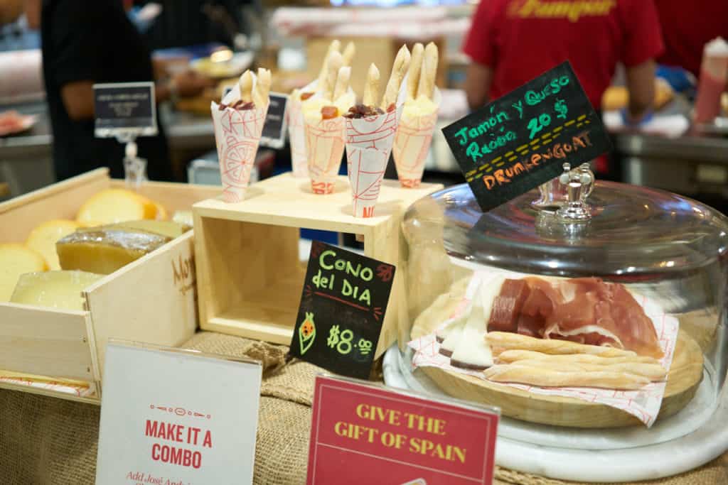 Cones with cheese, charcuterie and breadsticks on display next to the same ingredients under a glass dome.