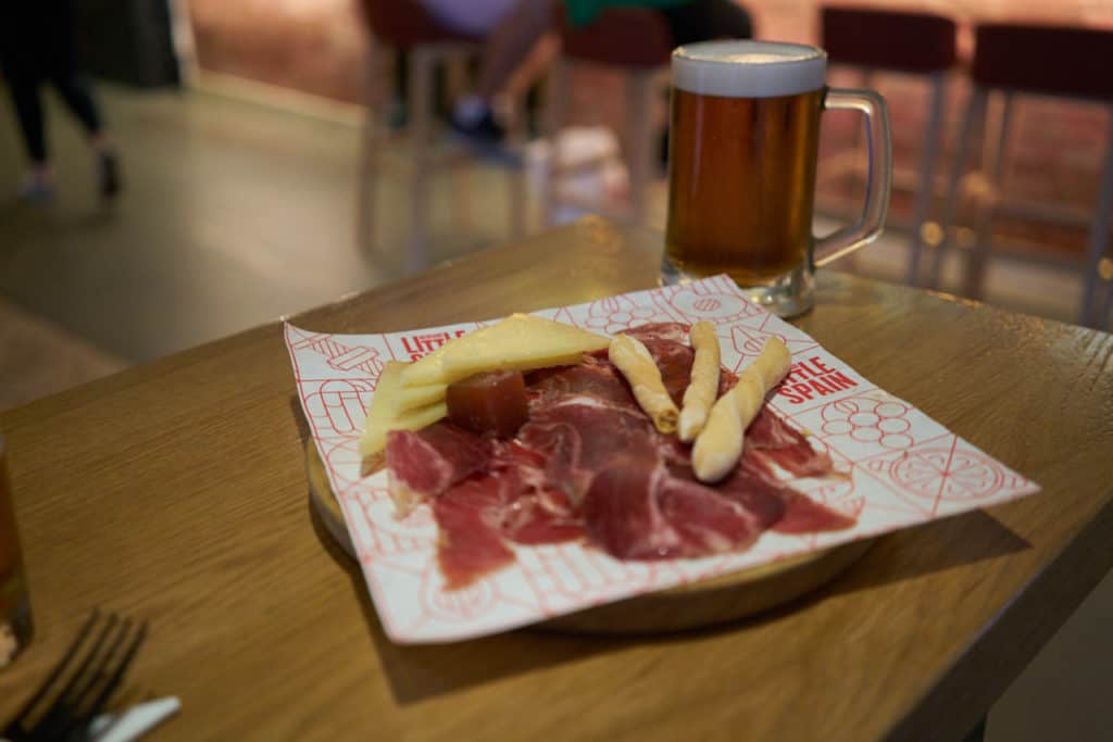 A plate of jamón iberico and manchego cheese with breadsticks on a table with a beer in the background.
