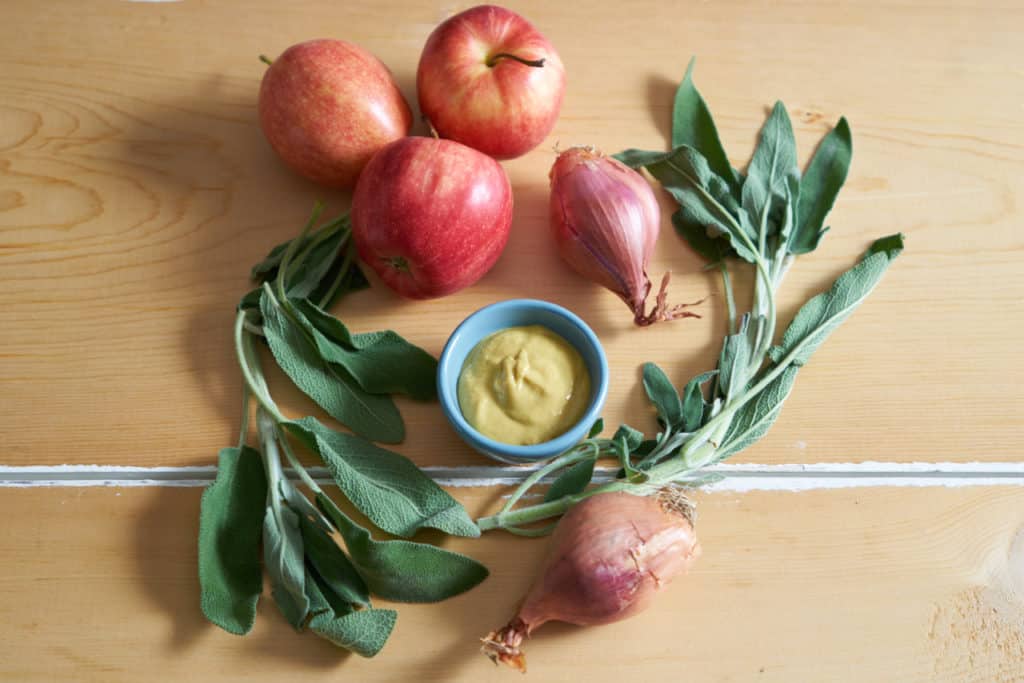 Sage, apples, shallots and a small dish of dijon mustard arranged on a wooden surface.