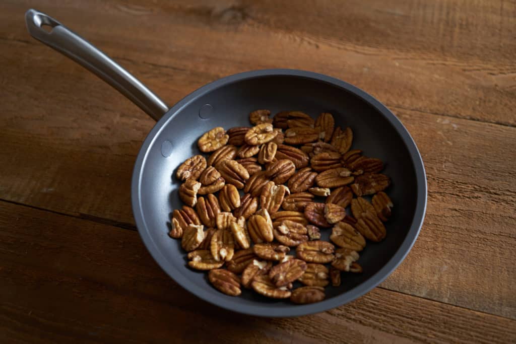 Pecan halves in a non stick skillet sitting on a wooden surface.