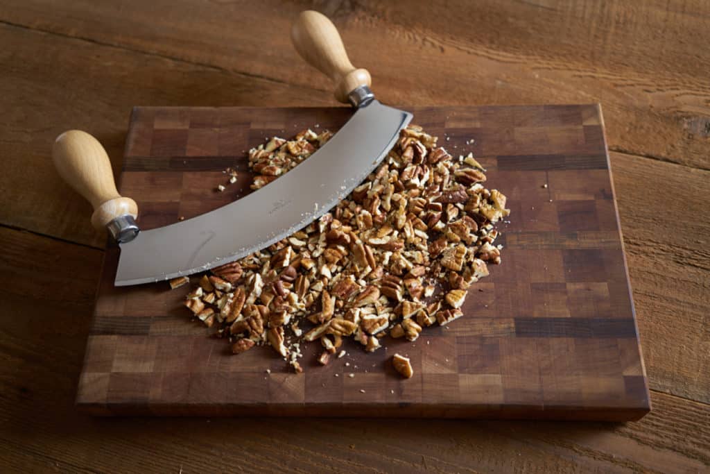 Chopped pecans and a mezzaluna on a wooden cutting board.