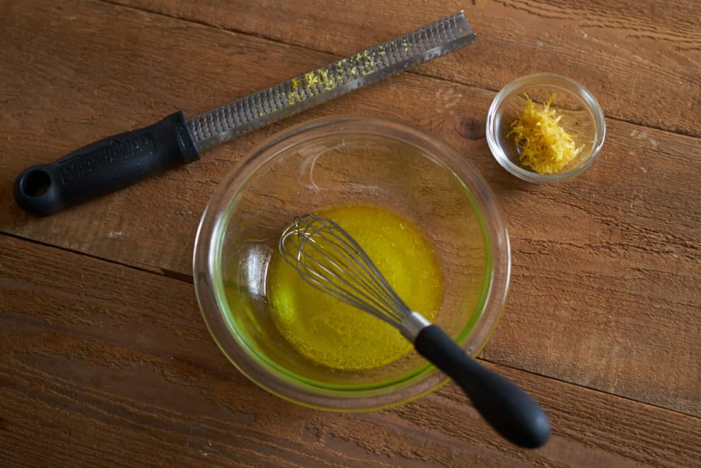 A glass bowl of lemon vinaigrette with a whisk in it. A zester and a small bowl of lemon zest sit on either side.