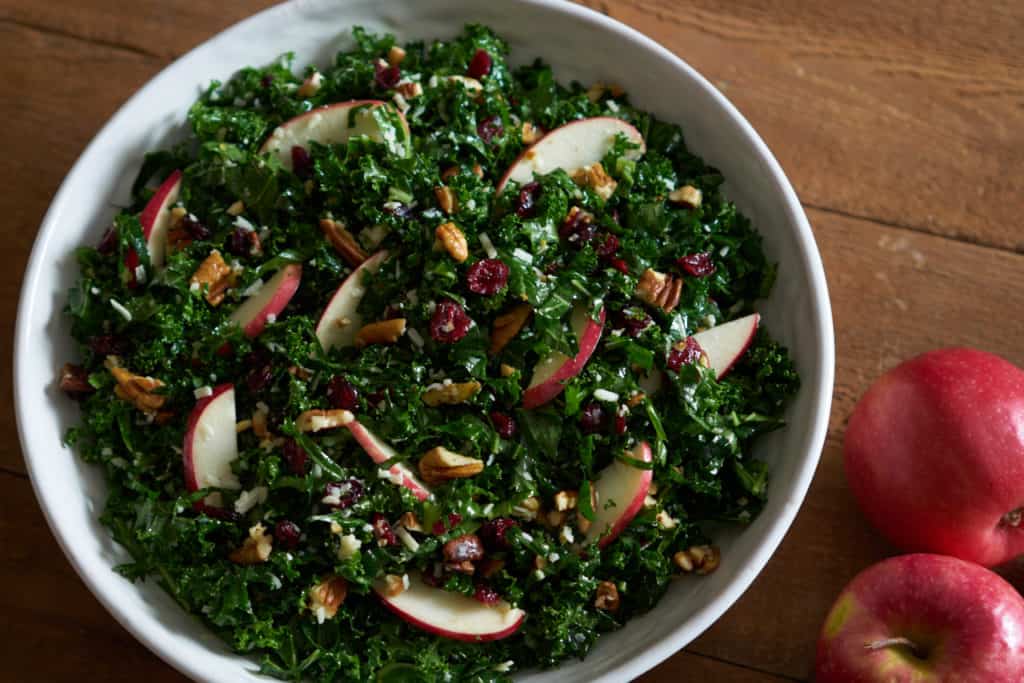 Kale salad with cranberries, apples and pecans in a white bowl on a wooden surface next to two red apples.
