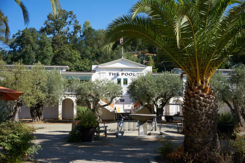 The Pool at Indian Springs, a white stucco building with an American flag on top, a fountain, palm trees and eucalyptus trees are in the foreground.