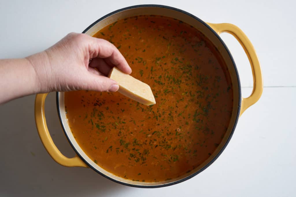 A woman's hand is shown adding a parmesan rind to a yellow pot filled with tuscan white bean soup.