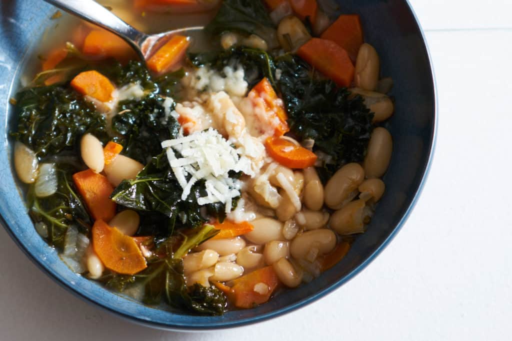 Tuscan White Bean Soup with Kale topped with parmesan cheese in a blue bowl with a spoon in it.