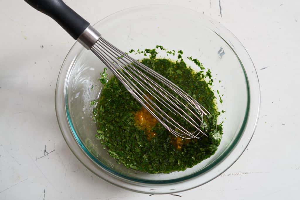A whisk sits inside a glass bowl filled with chopped herbs, olive oil and citrus zest.