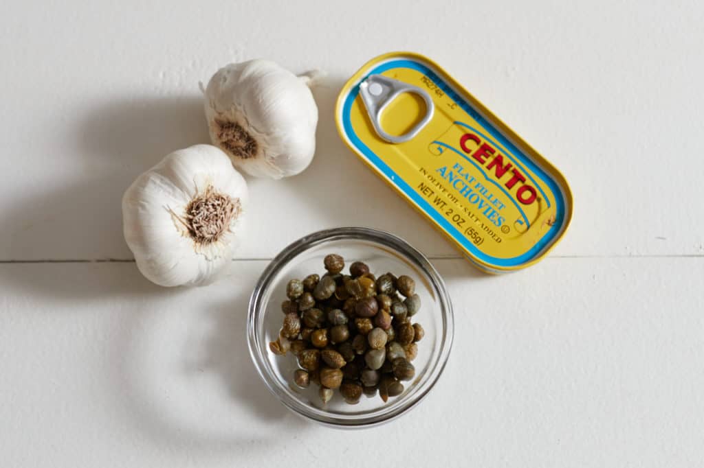 Capers in a small bowl, two cloves of garlic, and a tin of anchovies on a white surface.