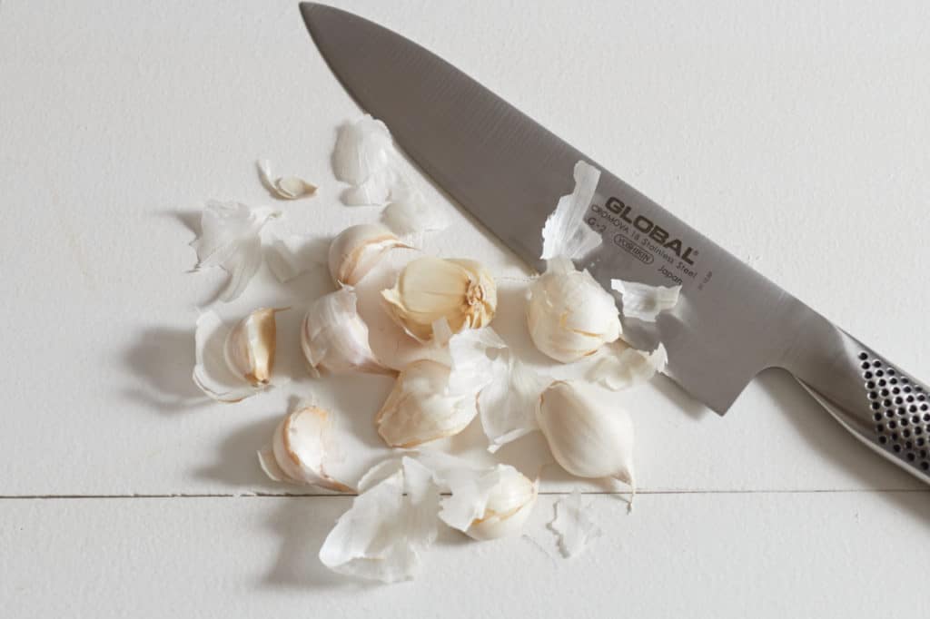 A chef's knife and several garlic cloves on a white surface.