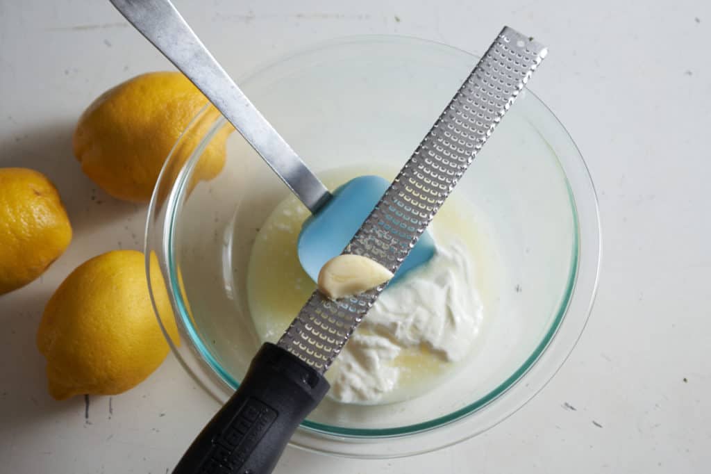 A microplane grater with a clove of garlic sits across glass bowl of yogurt and lemon juice.