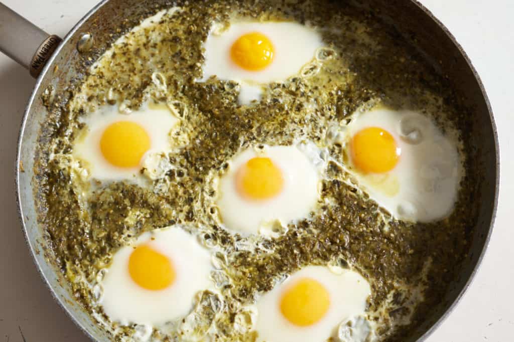 Six cooked sunny-side up eggs in green shakshuka sauce in a large skillet.