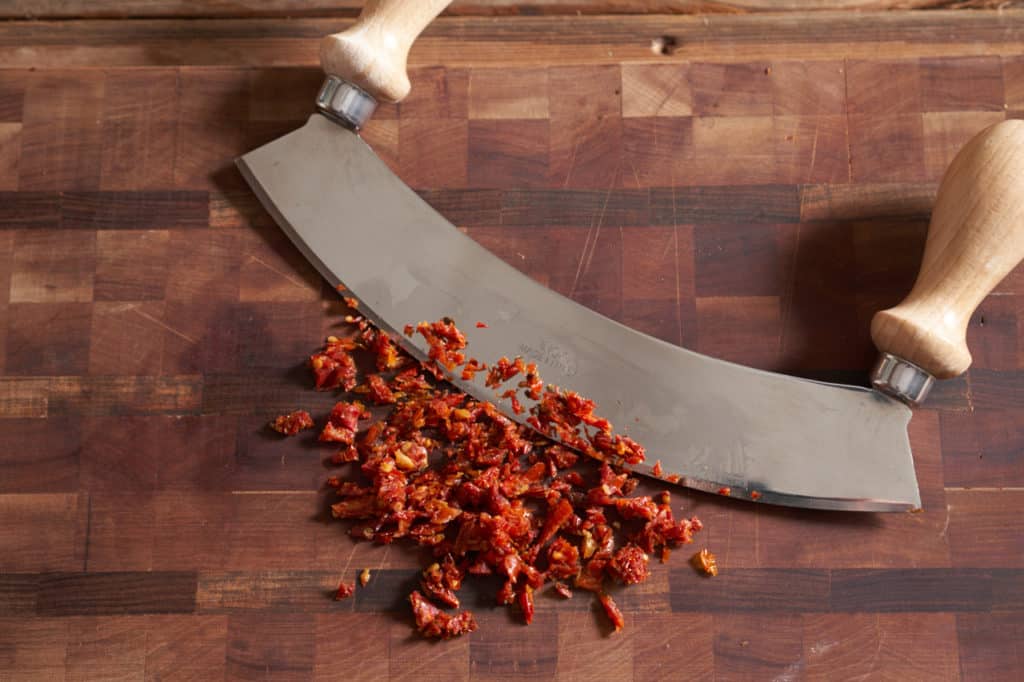 A mezzaluna on a cutting board with diced sun-dried tomatoes.