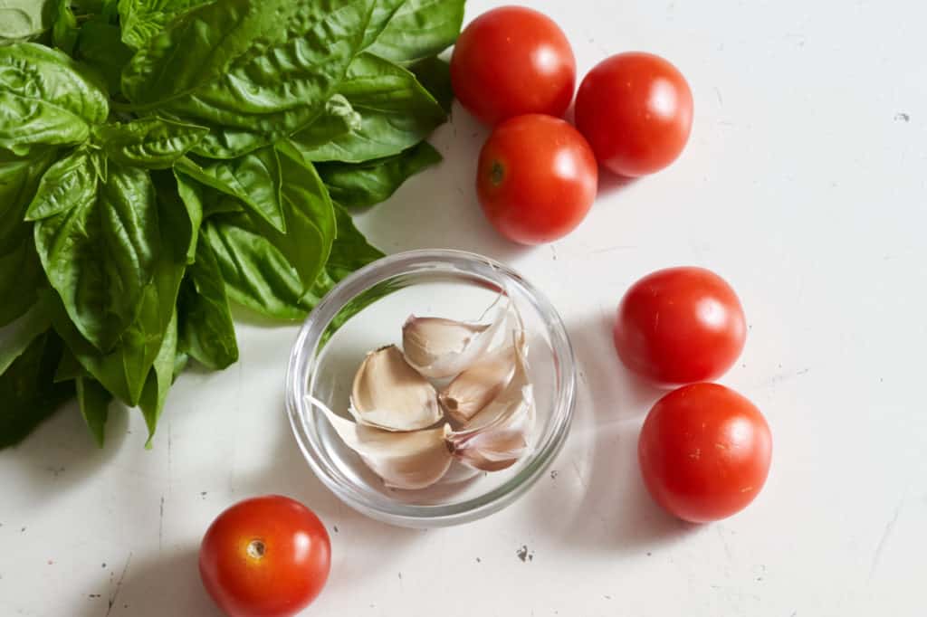 Fresh basil, cherry tomatoes and a small glass bowl filled with garlic cloves.