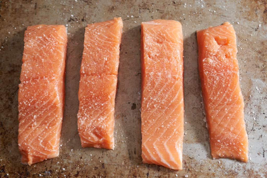 Four filets of raw salmon seasoned with salt on a baking sheet.