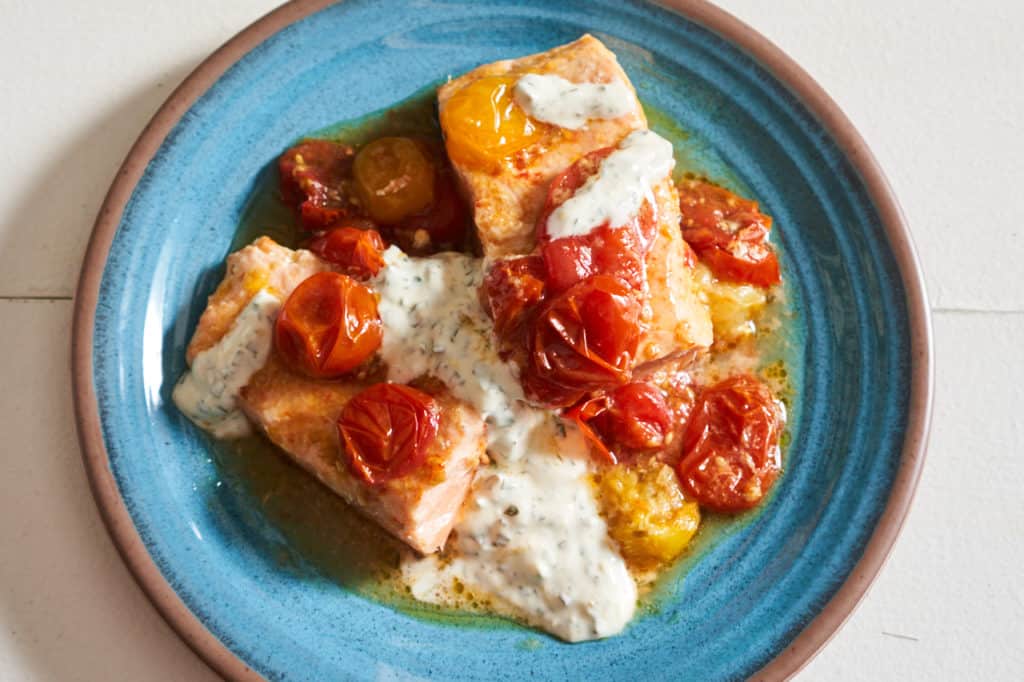 Oven baked salmon with roasted tomatoes and basil yogurt sauce on a blue plate.