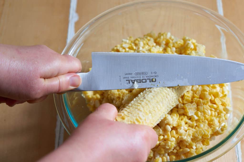 A woman's hands are shown using the back of a knife to scrape corn from a cob into a bowl.