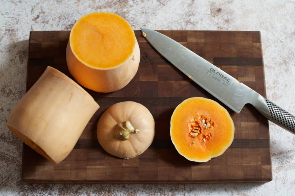 A butternut squash cut in half with the top and bottom cut off on a cutting board.