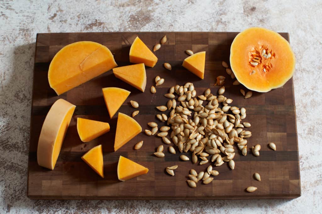 A butternut squash cut into triangle-shaped pieces on a cutting board along with its seeds.