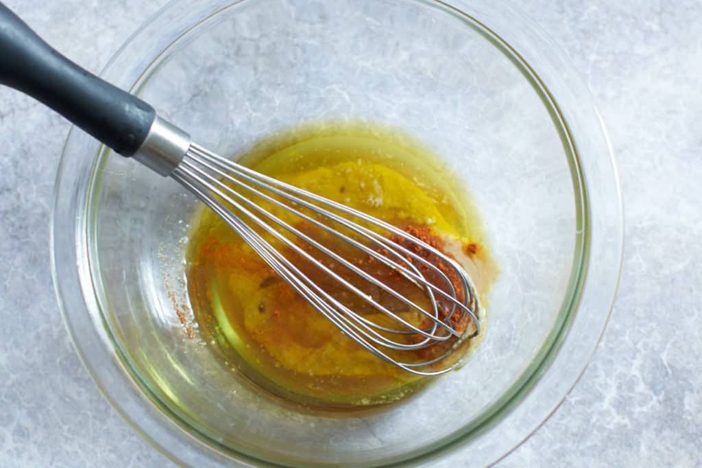 A whisk sits in a bowl of olive oil and spices.