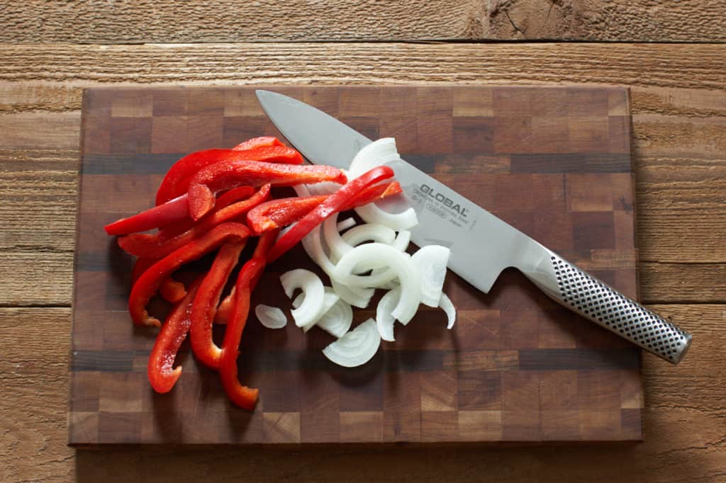 Sliced red bell peppers and onions on a cutting board with a knife.