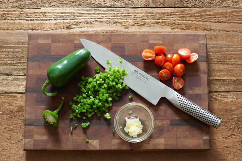 Chopped jalapeños, sliced cherry tomatoes and grated garlic on a cutting board with a knife.