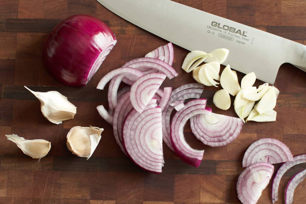 A chef's knife on a cutting board with slice red onion and garlic.