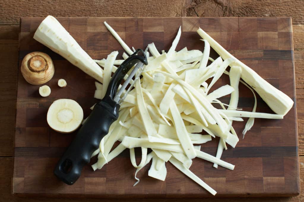 Shaved parsnips on a cutting board with a vegetable peeler.