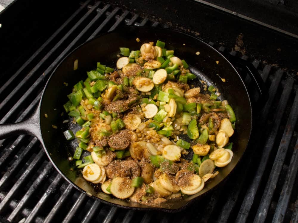 Potatoes and green peppers with taco seasoning cooking in a cast iron skillet on a grill. 
