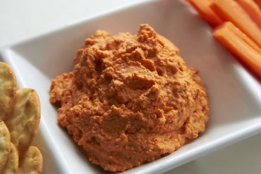 Creamy roasted red pepper dip in a white serving dish with carrot sticks and crackers on either side. 