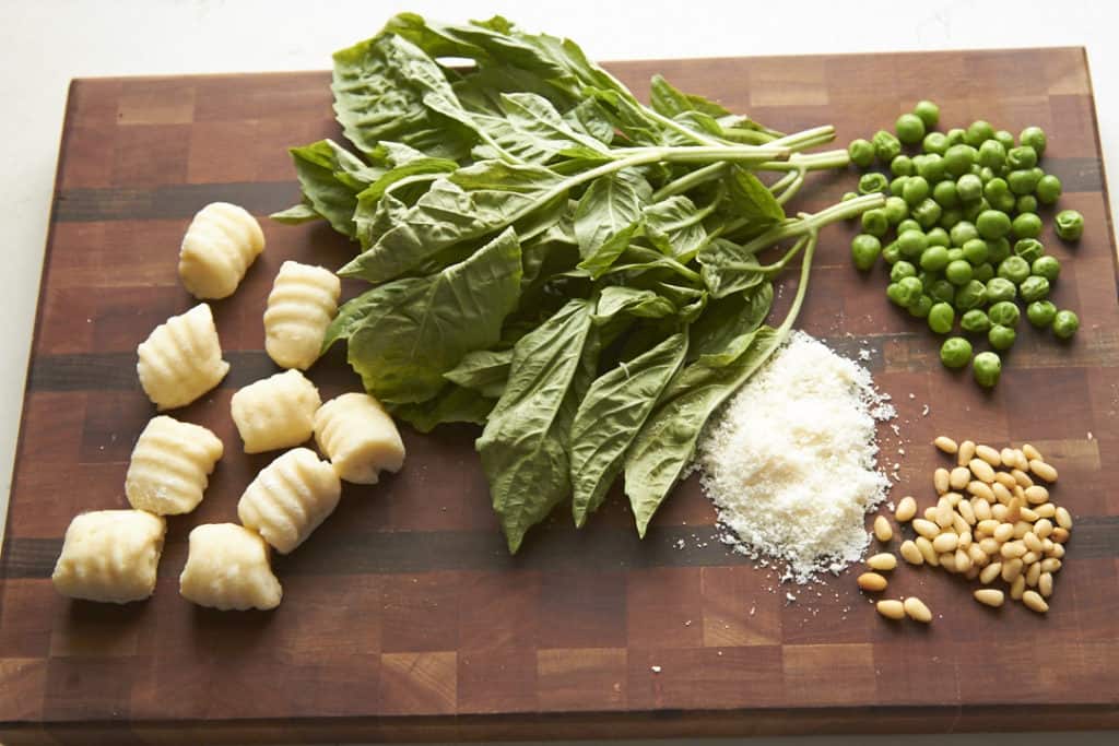 Gnocchi, fresh basil, peas, parmesan cheese, and pine nuts on a wooden cutting board.