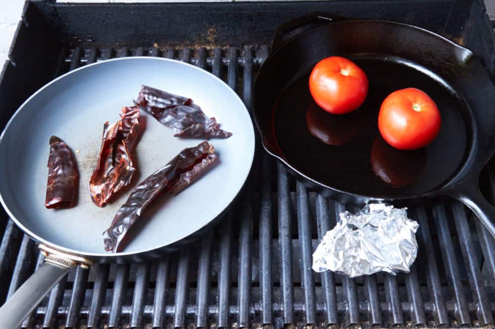 A grill with two skillets, one containing dried chiles, the other containing tomatoes. A foil pouch with roasting garlic sits beside them. 