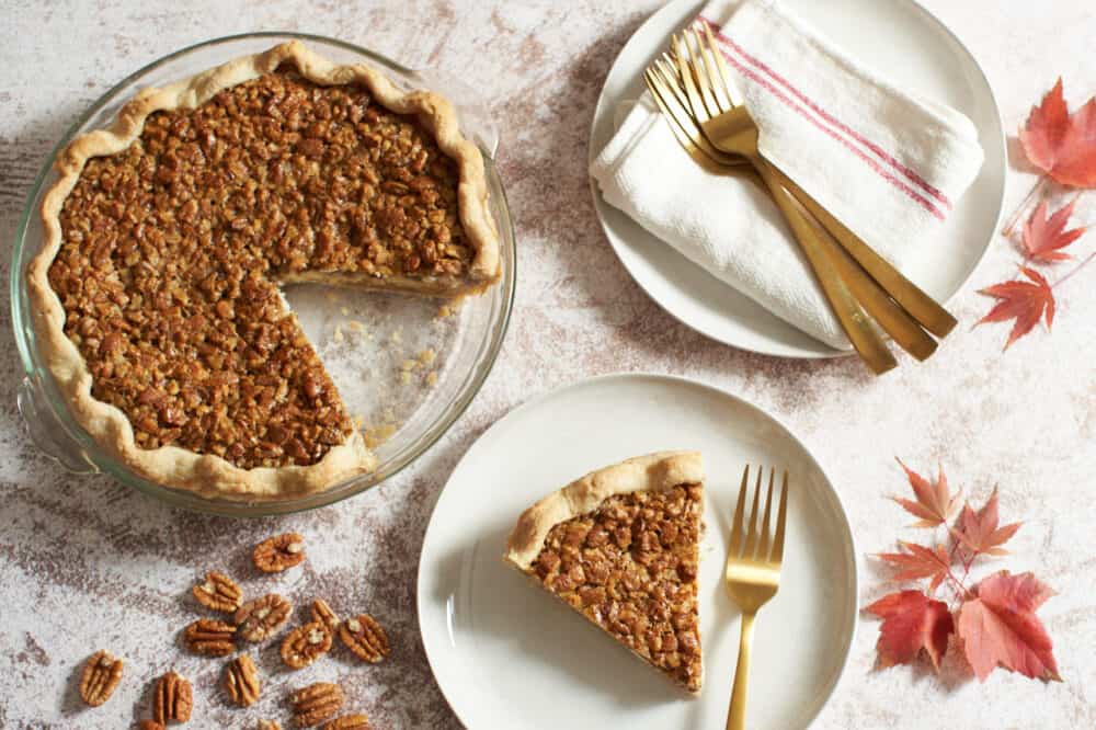 A pecan cream cheese pie with a slice cut out of it next to a white plate with a slice of pie on it. They are surrounded by more plates stacked with forks and napkins and some red leaves.