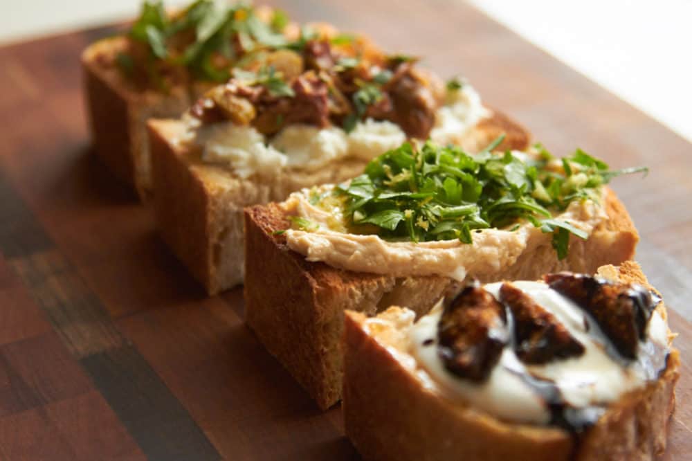 Four different bruschetta recipes displayed on a cutting board, with goat cheese, hummus, olive tapenade and other ingredients.