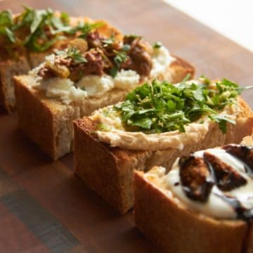 Four crostini with different toppings on a wooden cutting board.