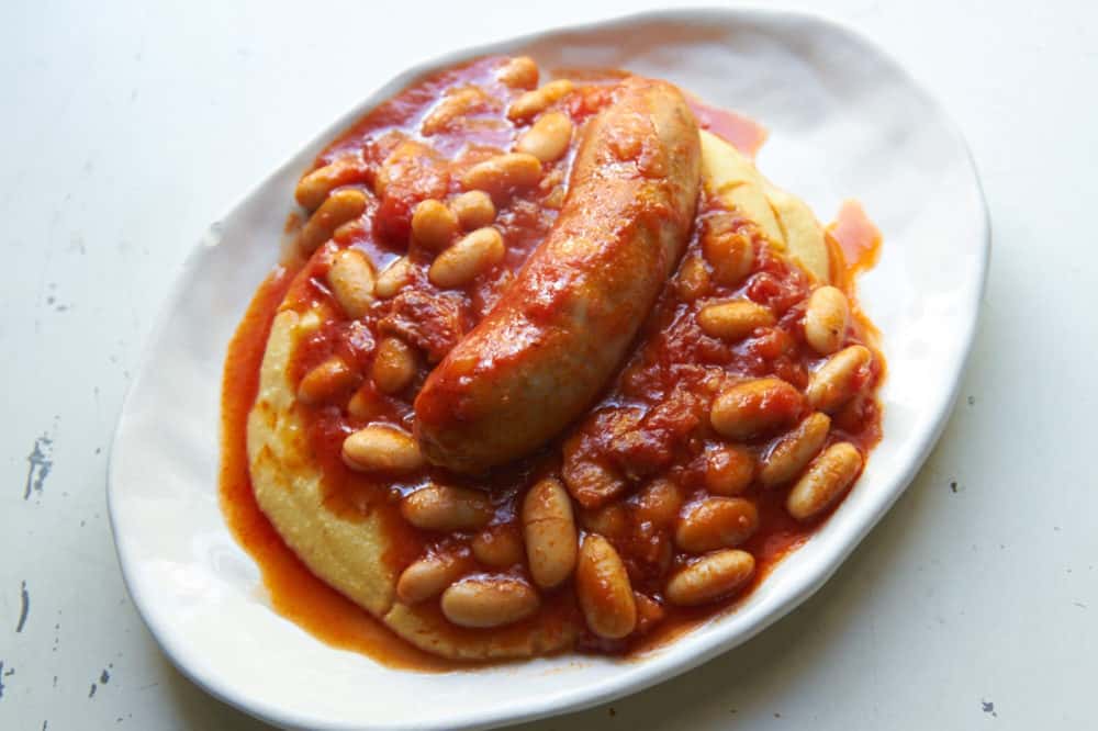 Sausage and cannelini beans in tomato sauce over polenta on a white plate. 