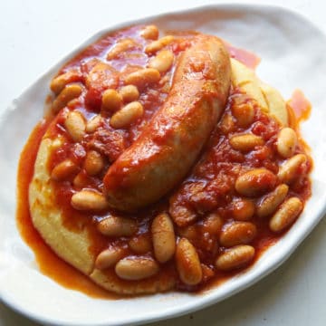 Italian sausage and cannelini beans in tomato sauce over creamy polenta on a white plate.