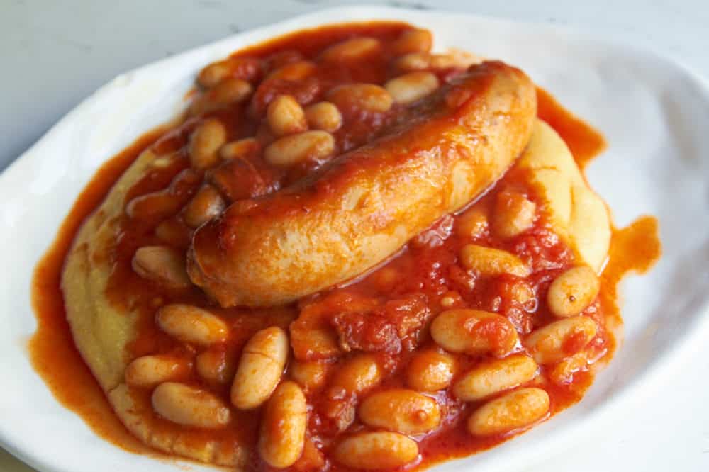Sausage and cannelini beans in tomato sauce over polenta on a white plate.